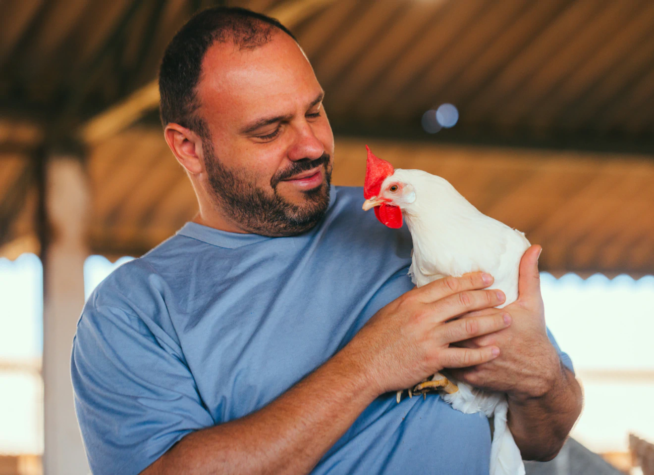 Homem sorridente segurando galinha branca com crista vermelha em seus braços dentro de aviário